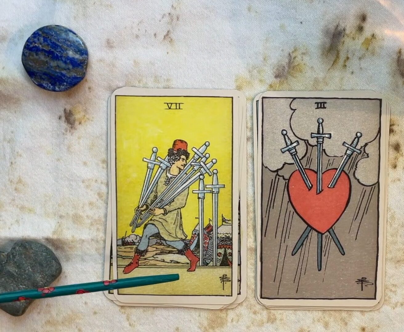 The seven of swords and the three of swords laid on a tarot reading space with decorative rocks.