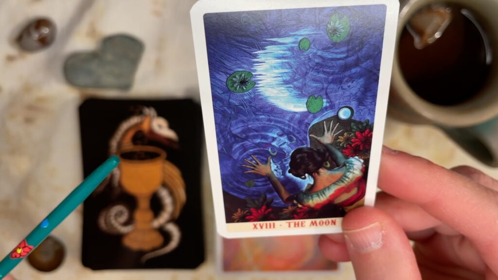 The Moon XVIII card from the Santa Muerte Tarot (mini), depicting a night scene, with a woman gazing at her reflection in a pond.