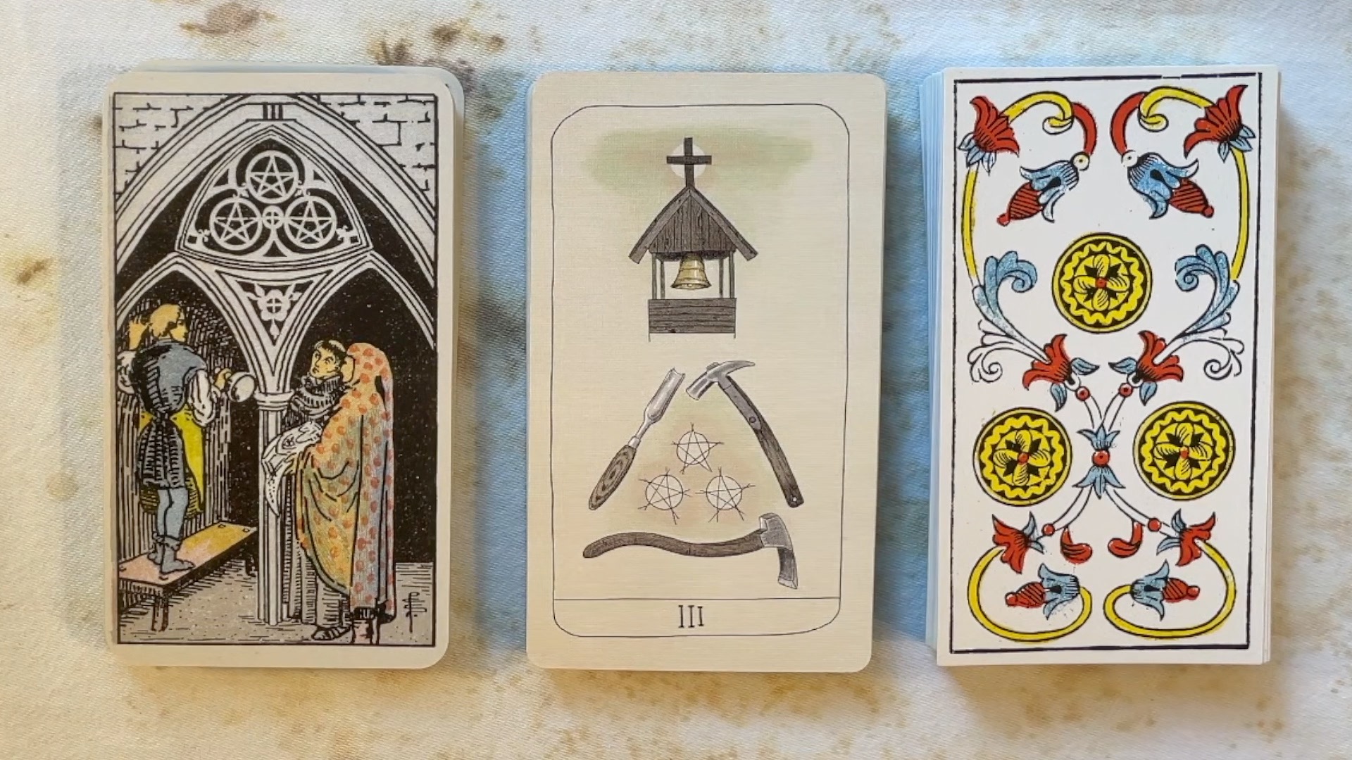 Three examples of the 3 of Pentacles / Coins: RWS on the left shows 3 figures standing under a church archway, the 5th Sprit in the center shows a church bell tower, worker implements, and three pentacles in the center, and the card on the right is a typical Marseille tarot showing three gold coins in the center, flanked by floral embellishments.